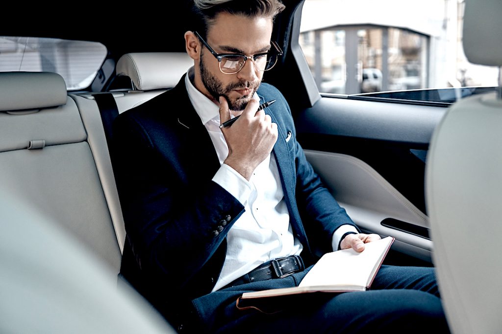 Thinking about business. Handsome young man in full suit writing something down in personal organizer while sitting inside of the car